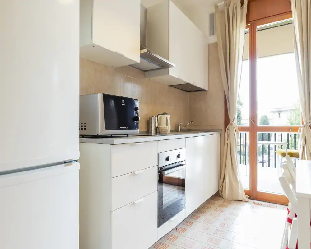 Bright modern kitchen with white cabinets, built-in oven, microwave and a large glass door opening to a balcony that provides generous natural light.