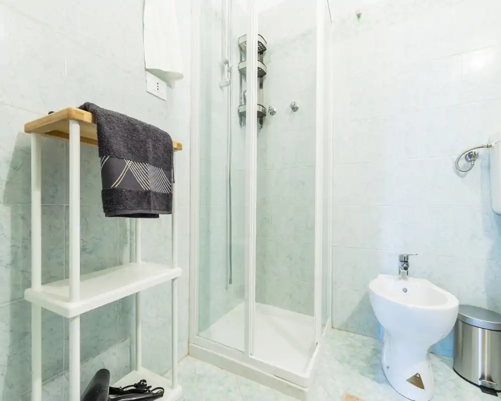 Clean, compact bathroom with a glass shower enclosure, bidet, and tidy shelving — bright mint tiles and modern fixtures create a fresh feel.