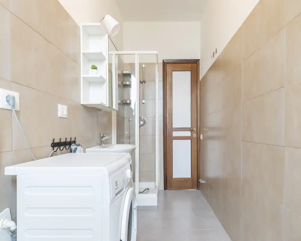 Compact, modern bathroom with a shower cabin, sink and a washing machine. Neutral beige tiles and clean lines create a bright, functional utility space.