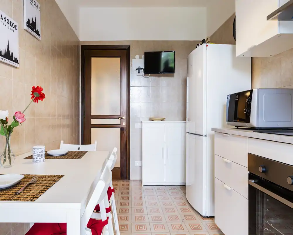 Bright, tidy kitchen with a dining table set for two, white cabinets, refrigerator and countertop appliances. Decorative flowers and placemats give a welcoming, lived-in feel.