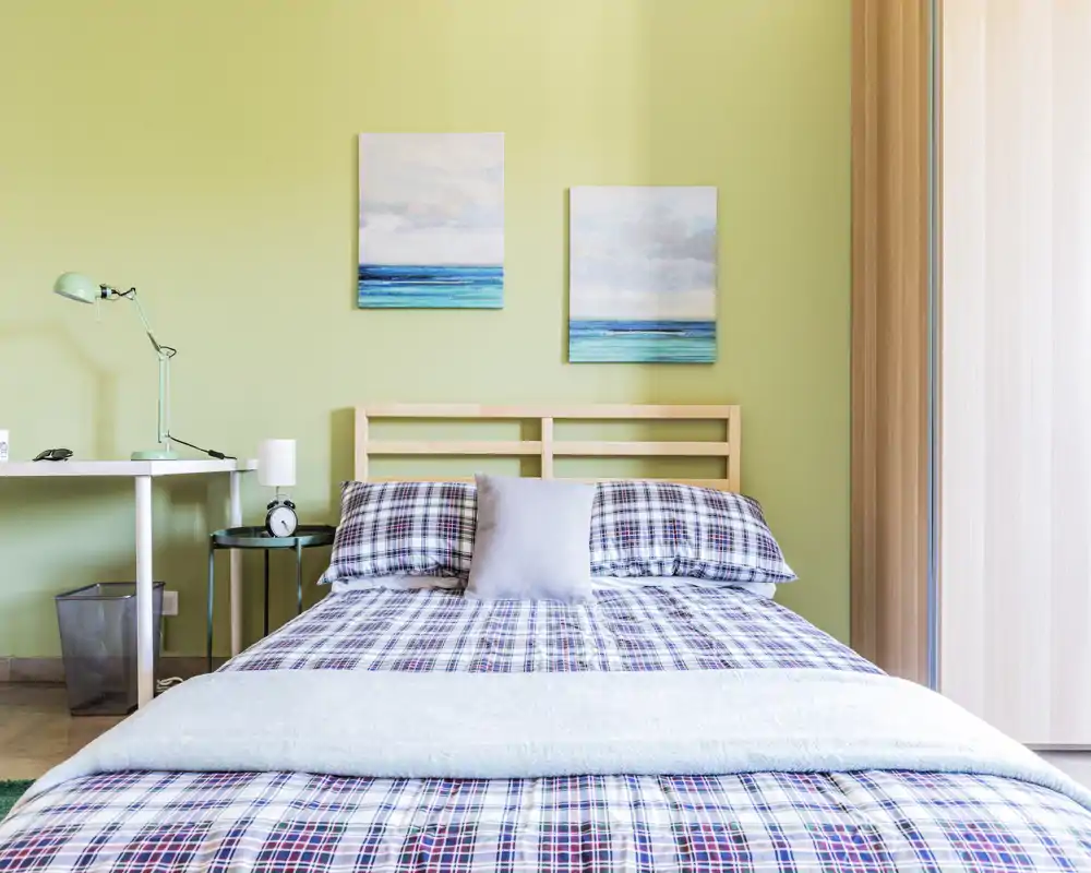 Dormitorio moderno y luminoso con cama doble bien hecha, ropa de cama de cuadros, dos cuadros con paisaje marino sobre la cabecera y mesita con lámpara — perfecto para un anuncio de alquiler.