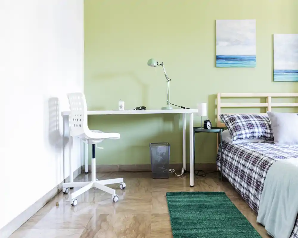 Dormitorio luminoso y sencillo con cabecero de madera, ropa de cama a cuadros, mesita con lámpara y despertador, y un escritorio ordenado con silla — ideal para una persona o estudiante.