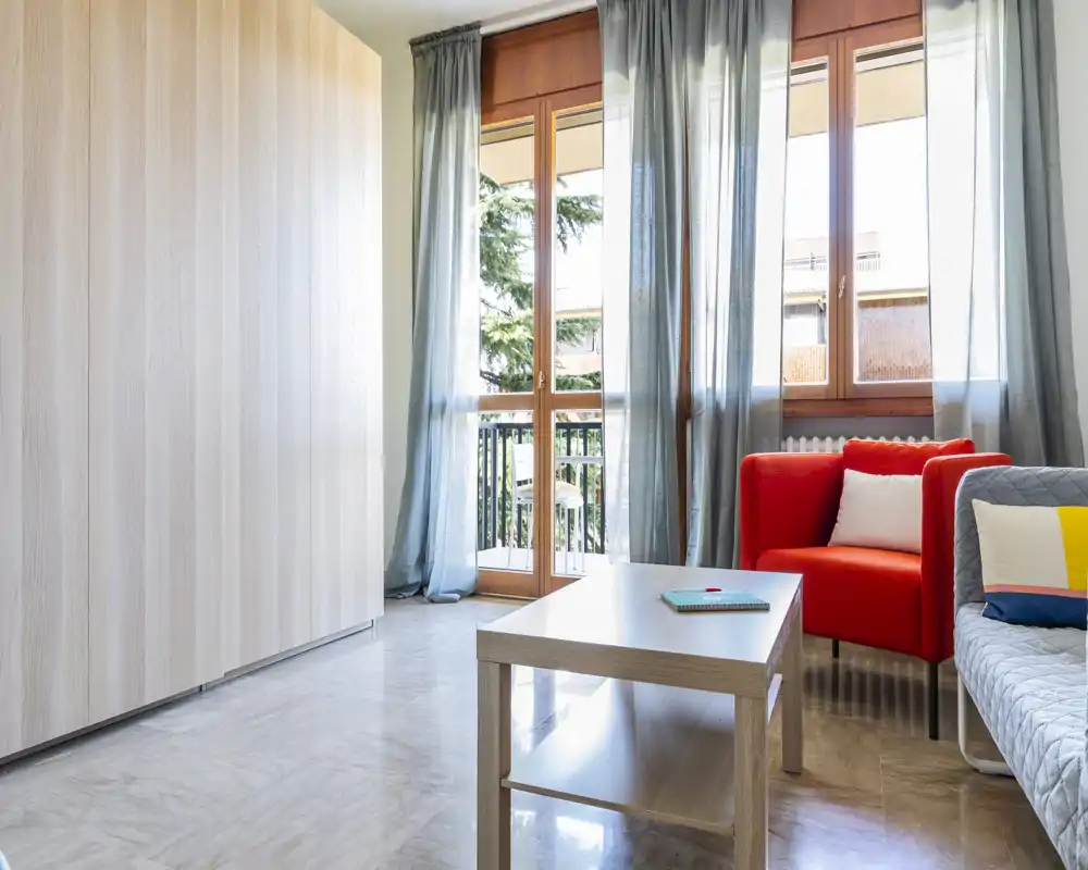 Zona de estar luminosa y moderna con grandes ventanales, un sillón rojo, sofá con cojines y una mesa de centro — ideal para relajarse con luz natural.
