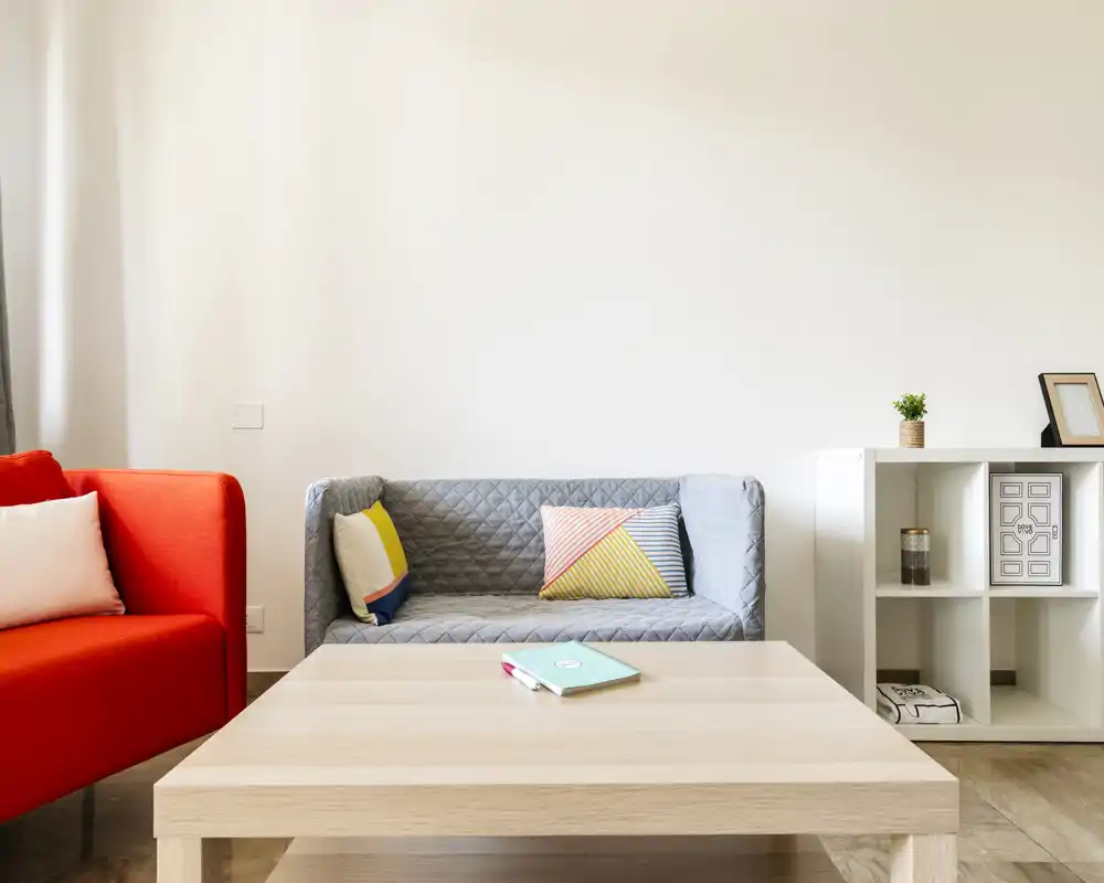 Sala de estar moderna y luminosa con sofá, sillón rojo y mesa de centro de madera clara — cojines y estantería minimalista crean un ambiente acogedor.