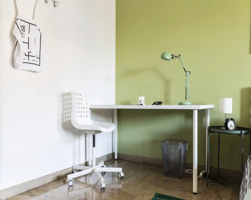 Zona de estar luminosa y minimalista con escritorio blanco y silla frente a una pared verde suave, dispuesta como un pequeño rincón de oficina en casa.
