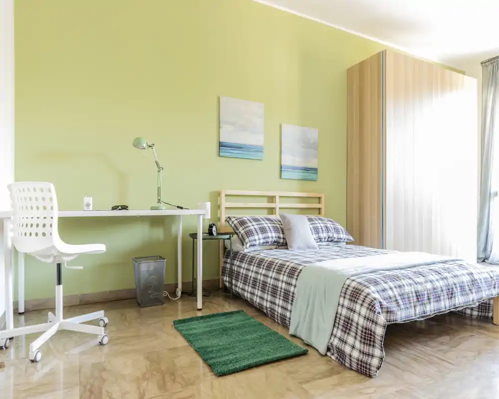 Dormitorio luminoso y bien decorado con cama doble, ropa de cama suave, mesita y un pequeño espacio de trabajo. La luz natural y los tonos verdes crean un ambiente relajante.