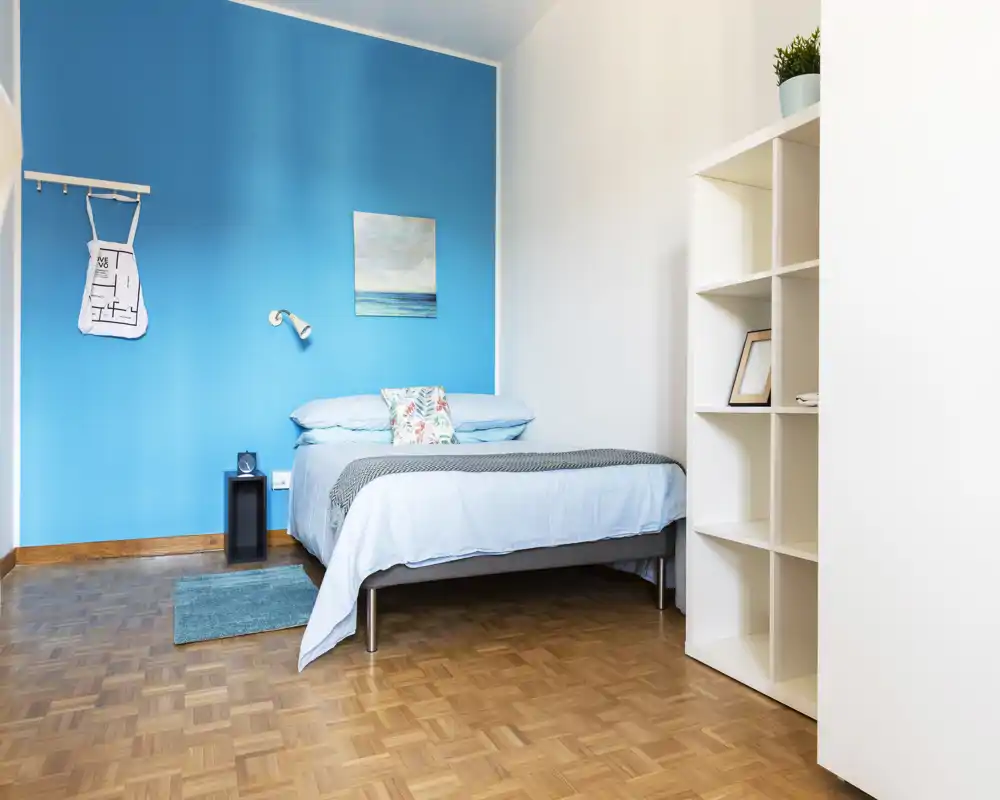 Dormitorio moderno y luminoso con pared turquesa, cama bien tendida, estantería sencilla y suelo de parquet — ideal para alquileres de corta estancia.