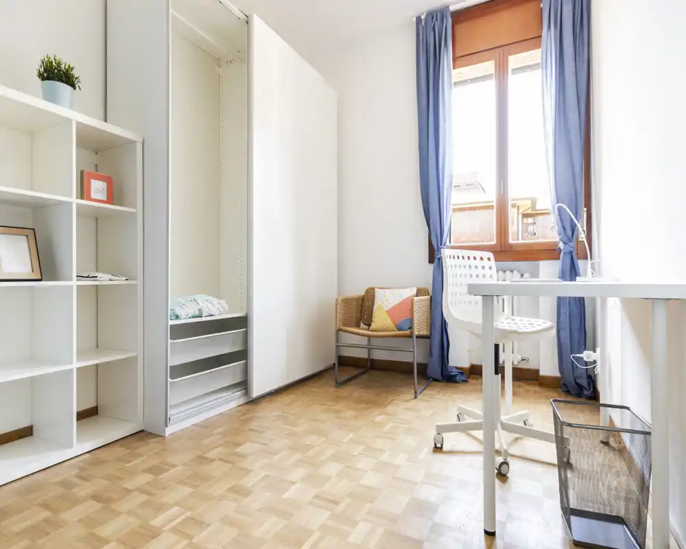 Habitación luminosa y minimalista con ventana, escritorio y estanterías: espacio versátil apto como dormitorio pequeño o estudio. Suelo de parquet, butaca de mimbre con cojín y muebles blancos ofrecen una sensación fresca y aireada.