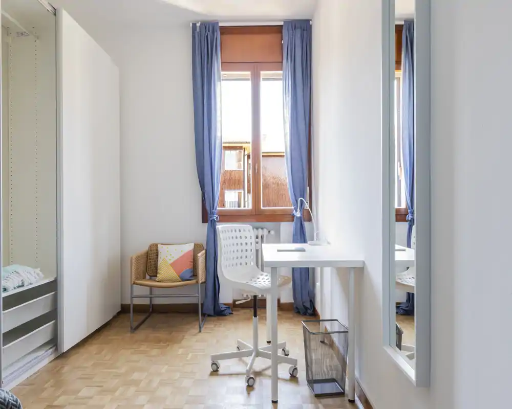 Rincón de dormitorio luminoso y minimalista con escritorio, silla y armario; luz natural desde la ventana y mobiliario moderno sencillo.