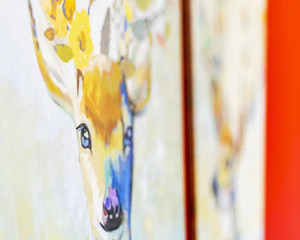 Primo piano di un dipinto o stampa decorativa con un cervo: colori vivaci (giallo, blu) su sfondo arancione, messa a fuoco ridotta per effetto bokeh.