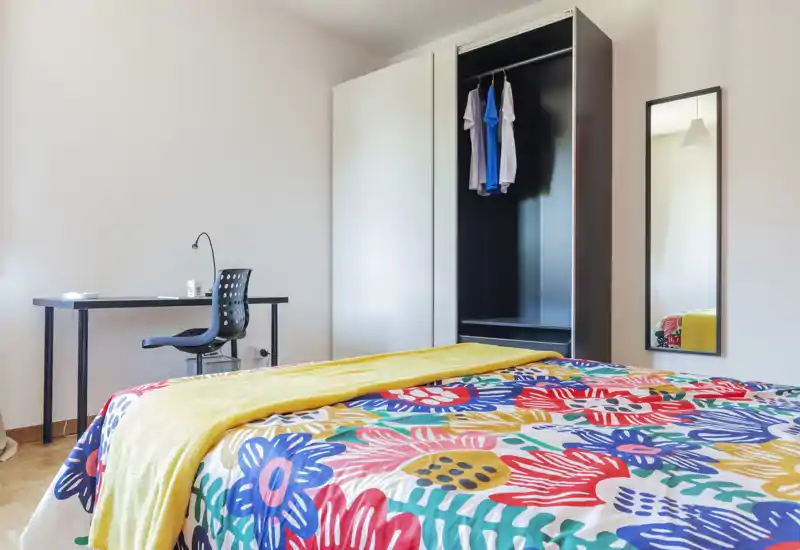 Camera da letto luminosa e moderna con copriletto floreale colorato, plaid giallo e un armadio con spazio appendiabiti. La stanza comprende una scrivania con sedia, uno specchio a figura intera e pavimento in parquet pulito: fresca, ben illuminata e accogliente.