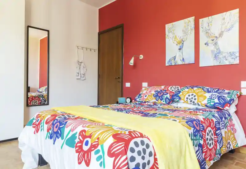 Camera da letto luminosa e colorata con un ampio letto rivestito da una biancheria floreale vivace su una parete rossa; due stampe con cervi e uno specchio a figura intera completano l'atmosfera accogliente e ben illuminata.