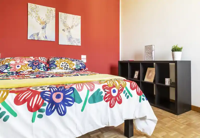 Camera da letto luminosa e colorata con copriletto floreale, cuscini decorativi e quadri coordinati; mobile a cubi moderno e pavimento in parquet per un ambiente accogliente.