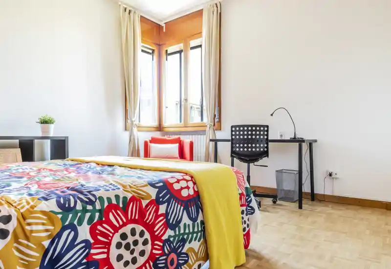Camera da letto luminosa e colorata con una grande copertura floreale, un plaid giallo e luce naturale dalla finestra angolare. Una scrivania e una sedia sul lato rendono la stanza adatta anche allo studio.