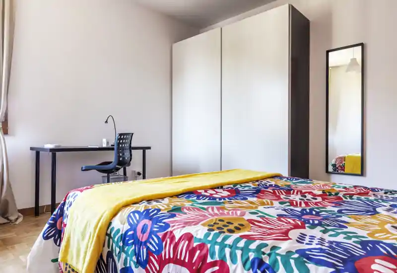 Camera da letto luminosa e ben arredata con una trapunta colorata, plaid giallo e una scrivania semplice vicino alla finestra — uno spazio accogliente e ordinato con luce naturale.