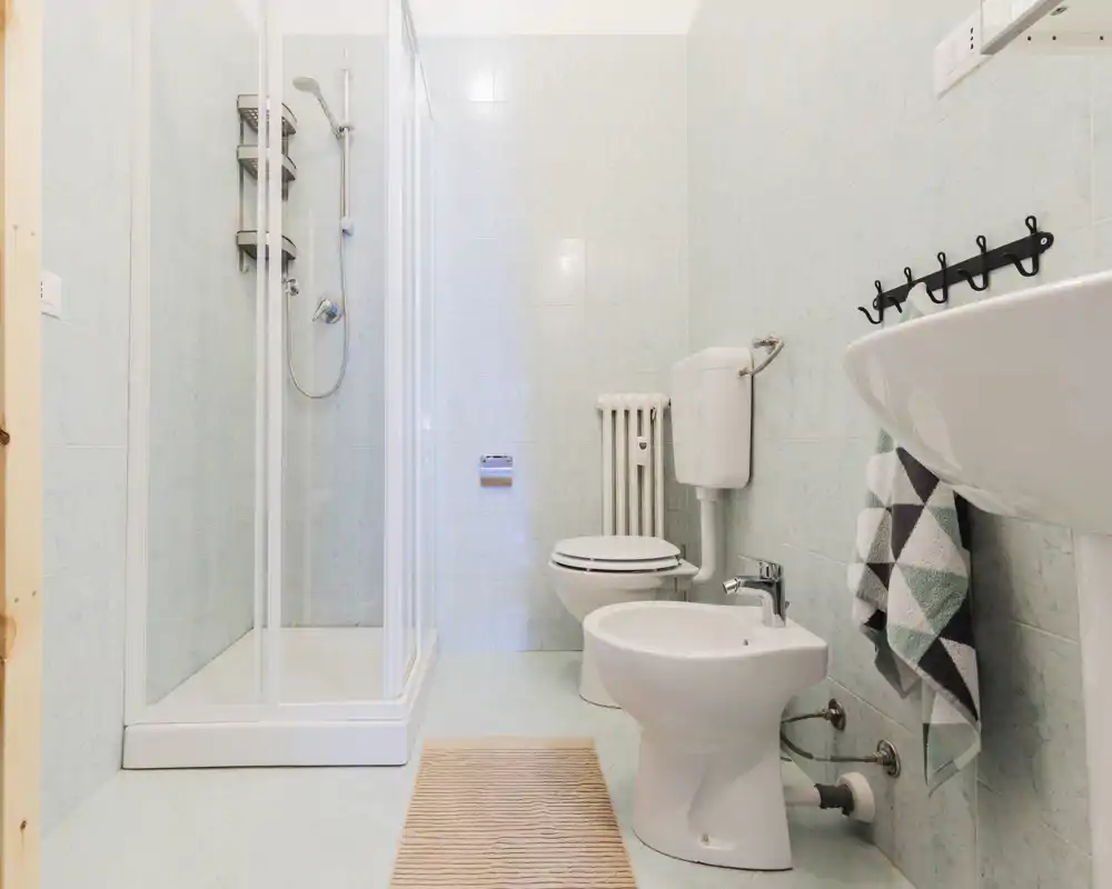 Bagno luminoso e pulito con box doccia, WC, bidet e lavabo a colonna; piastrelle chiare e illuminazione soffusa creano un ambiente fresco e arioso.