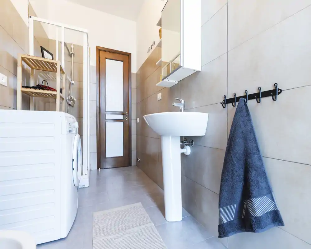 Bagno moderno e pulito con lavabo a colonna, specchio con pensile, box doccia e lavatrice; piastrelle neutre e un asciugamano appeso creano uno spazio ordinato e funzionale.