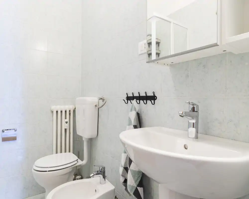 Bagno pulito e luminoso con lavabo a colonna, bidet, WC e specchiera; piastrelle chiare e rubinetteria cromata creano un ambiente funzionale e arioso.