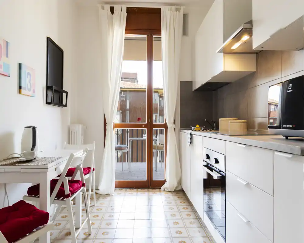 Cucina moderna e luminosa con mobili bianchi, forno integrato e un piccolo angolo pranzo accanto alla porta del balcone che illumina l'ambiente con luce naturale.