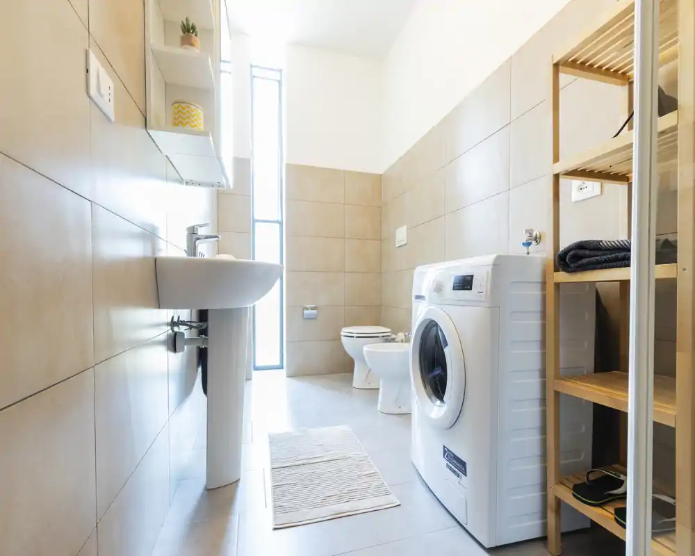 Bagno moderno e luminoso con lavabo a colonna, WC, bidet e una lavatrice a carica frontale—piastrelle pulite e luce naturale da una finestra stretta.