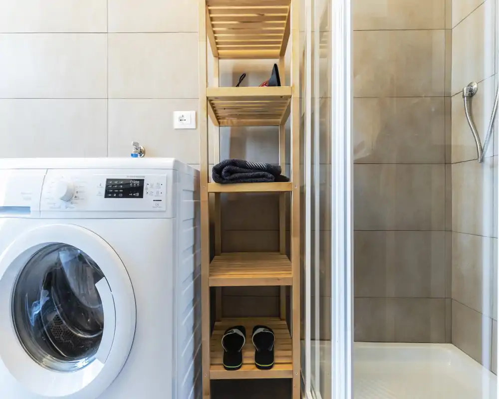 Bagno compatto con area lavanderia integrata: lavatrice a carica frontale accanto a scaffalatura in legno e box doccia piastrellato.