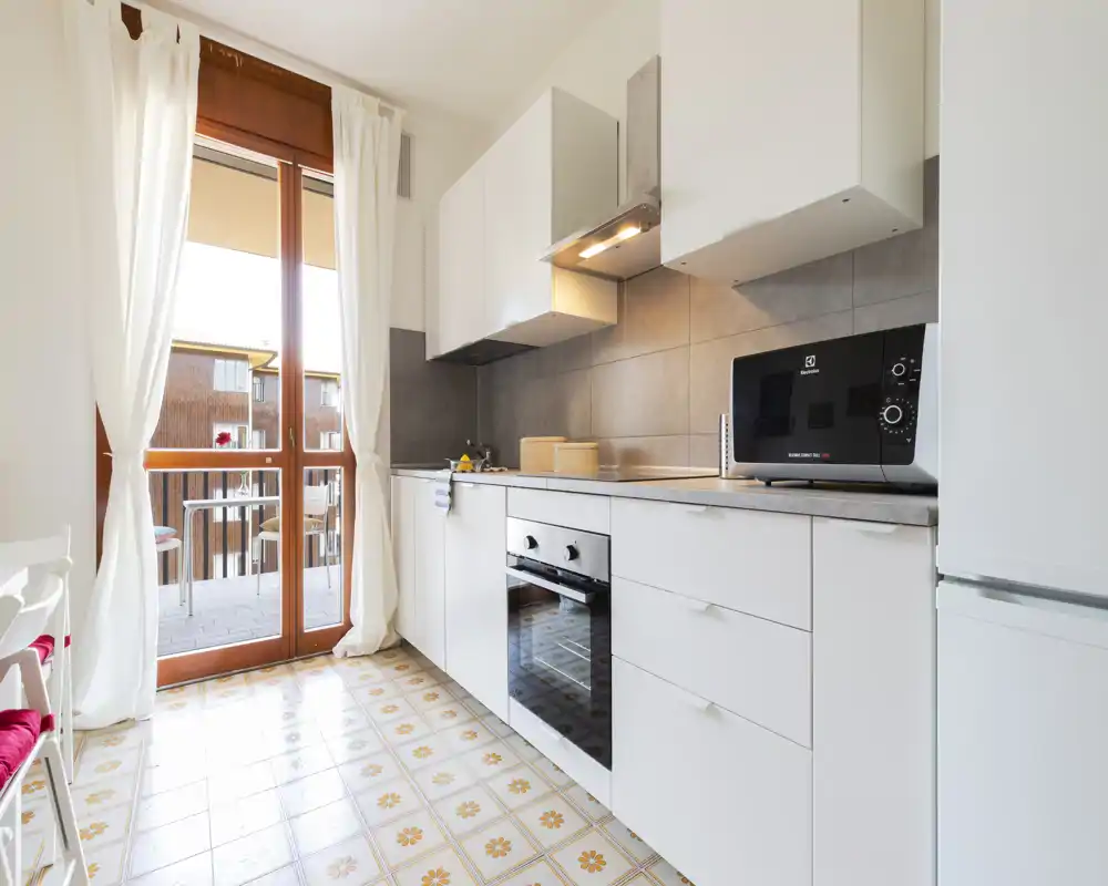 Cucina moderna e luminosa con forno incassato, microonde sul piano, mobili bianchi e grandi porte finestra che fanno entrare molta luce naturale.