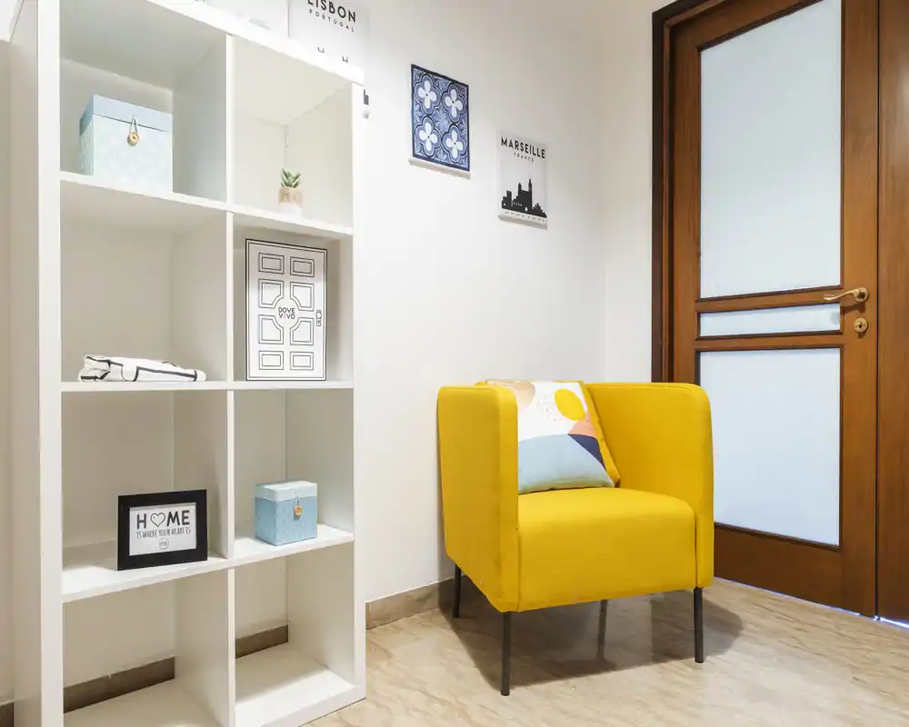Ingresso luminoso e ordinato con una poltrona gialla moderna, mensole decorative e una porta in legno con vetro satinato—ideale come accogliente disimpegno o angolo di ricevimento.