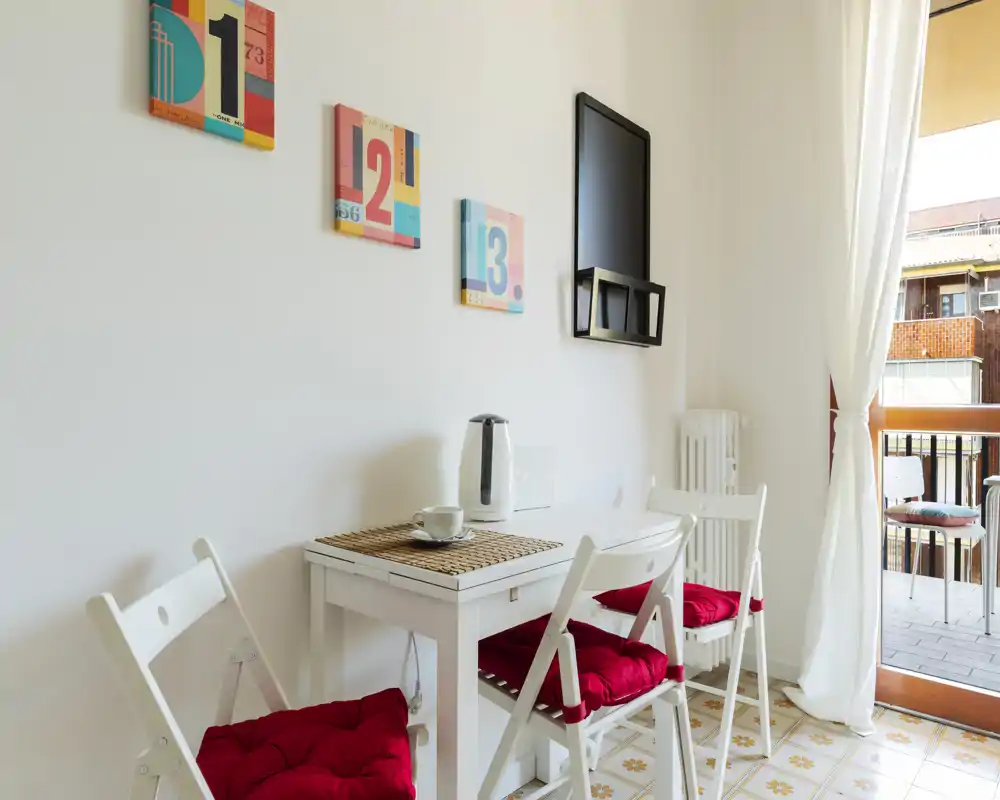 Angolo pranzo luminoso e compatto con tavolo bianco e sedie pieghevoli con cuscini rossi, quadretti colorati e luce naturale dal balcone — perfetto per una colazione accogliente.
