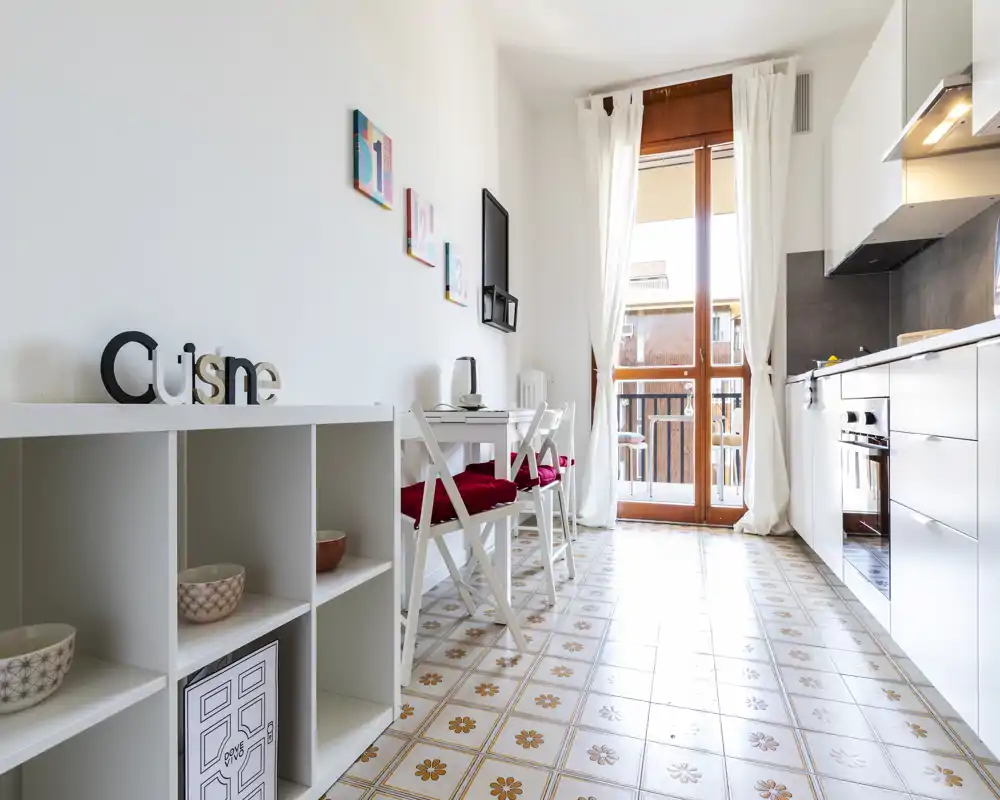 Angolo cottura moderno e luminoso con mobili bianchi, forno incassato e un piccolo spazio pranzo davanti a una grande porta finestra che dà sul balcone. Le piastrelle decorate e i cuscini rossi rendono l'ambiente accogliente.