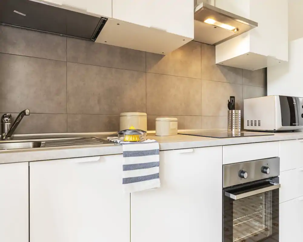 Cucina moderna e compatta con mobili bianchi, lavello in acciaio e forno integrato. Piano di lavoro pulito con barattoli, un asciugamano a righe e luce calda sotto il pensile, ambiente funzionale e accogliente.
