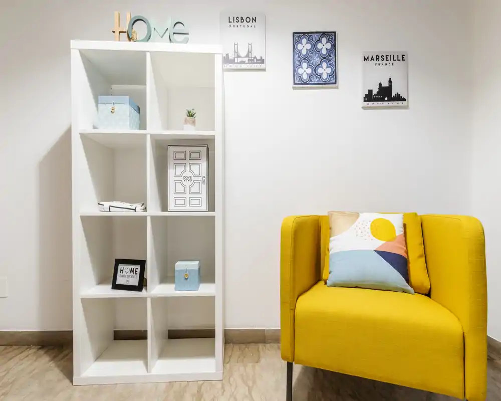 Angolo living luminoso e moderno con una poltrona gialla vivace, cuscino decorativo e una libreria bianca minimalista con quadri a parete — un angolo accogliente perfetto per rilassarsi o leggere.
