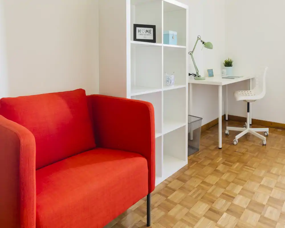 Pequeña zona de estar luminosa con un sofá rojo intenso, estantería abierta y un escritorio blanco compacto: un rincón moderno y acogedor ideal para relajarse o trabajar.