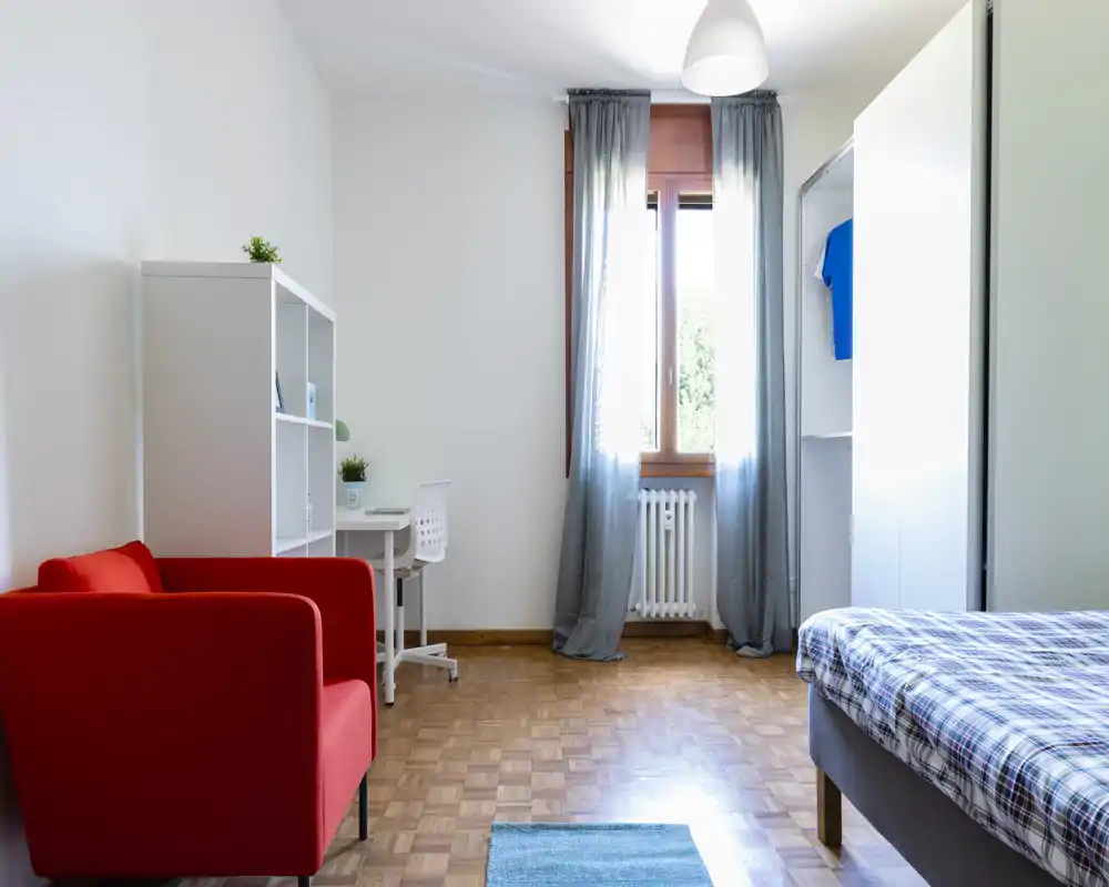 Dormitorio luminoso y ordenado con cama individual, sillón rojo, escritorio y estantería junto a la ventana. La habitación recibe buena luz natural y está amueblada con estilo sencillo y moderno.