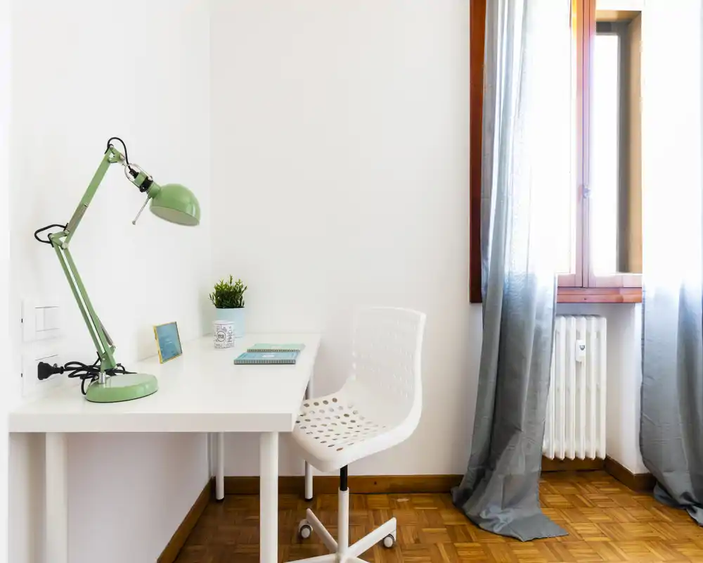 Rincón de trabajo luminoso y minimalista con escritorio blanco, silla perforada y una lámpara verde regulable junto a una ventana soleada: un espacio de trabajo ordenado y funcional.