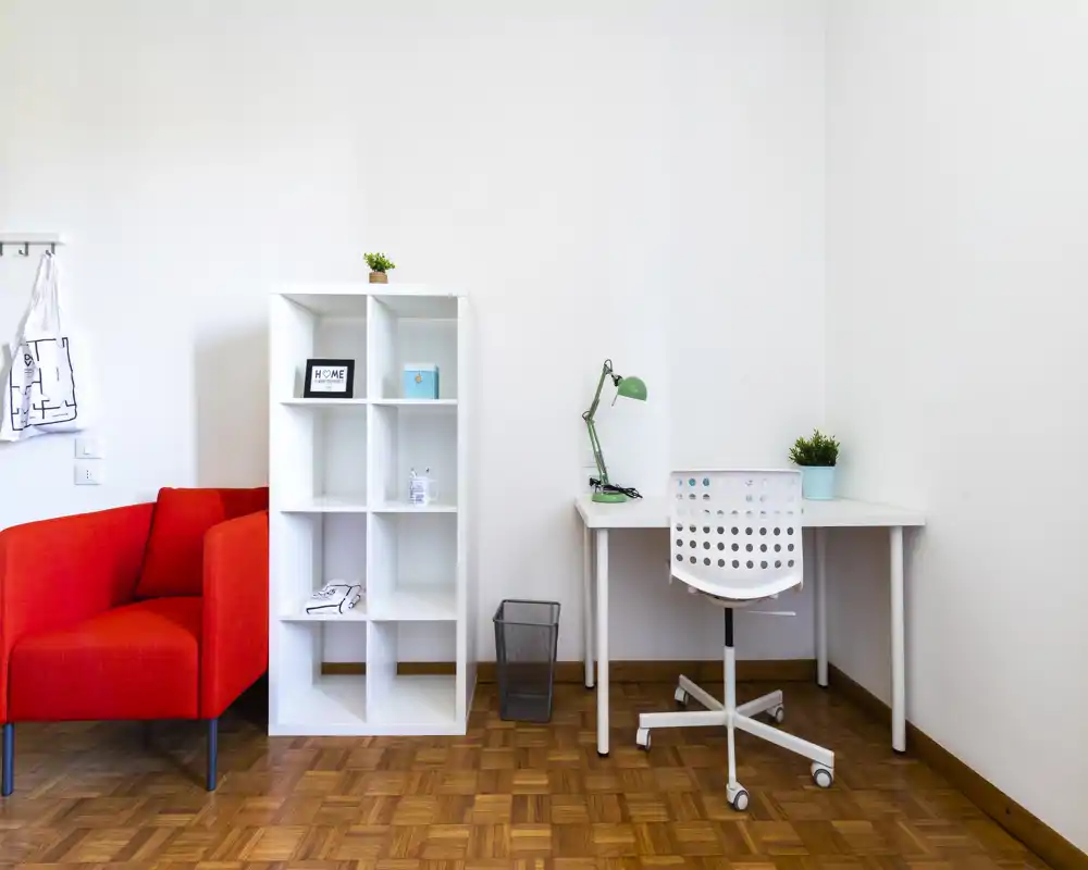 Zona de estar luminosa y minimalista con un sillón rojo, estantería blanca y un escritorio compacto — ideal para relajarse o trabajar en un apartamento pequeño.