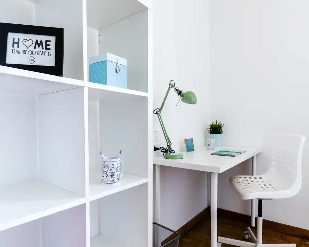 Espacio de trabajo luminoso y minimalista con escritorio blanco, silla perforada y estantería. Una lámpara verde, una planta pequeña y accesorios ordenados crean un rincón de estudio moderno.