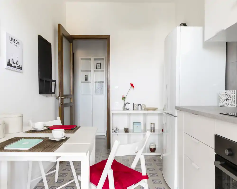 Cocina compacta y luminosa con pequeña mesa comedor, mobiliario blanco, frigorífico y detalles decorativos que crean un ambiente moderno y acogedor.