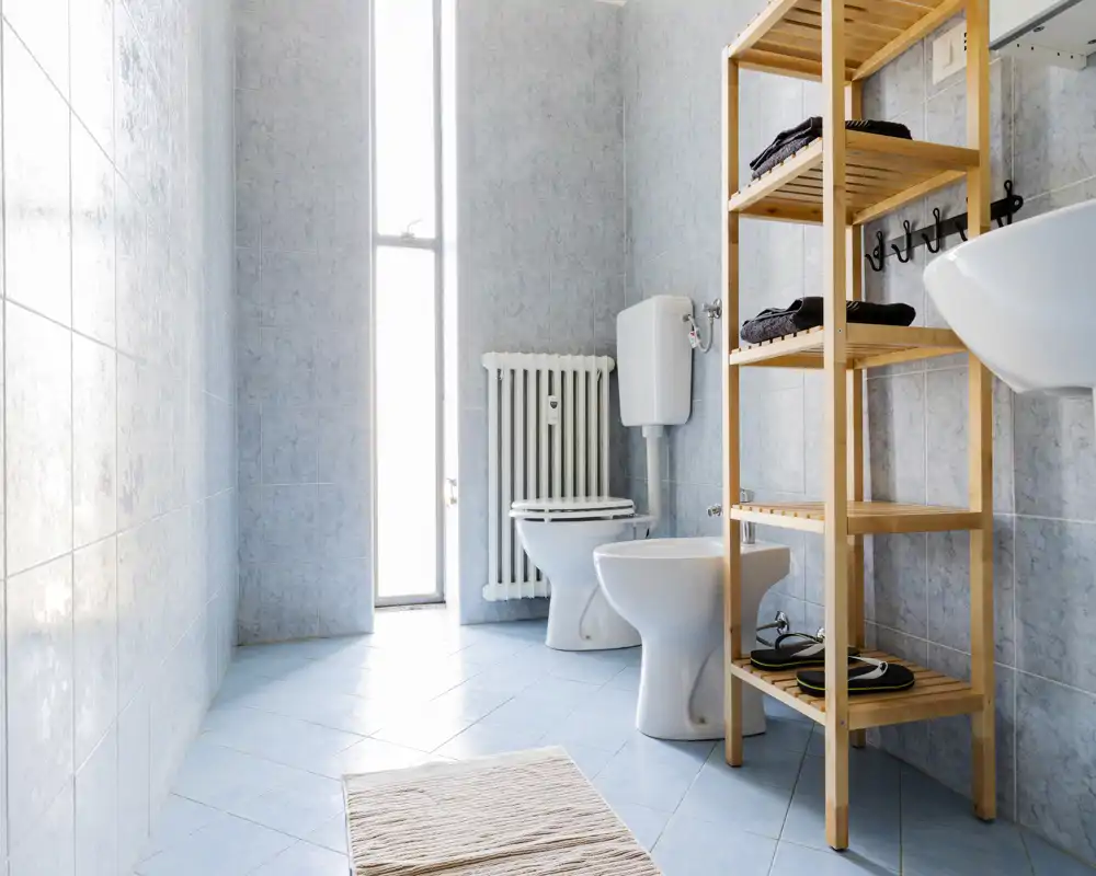 Baño luminoso y limpio con paredes y suelo alicatados, inodoro, bidé, lavabo y una estantería de madera para toallas y accesorios.