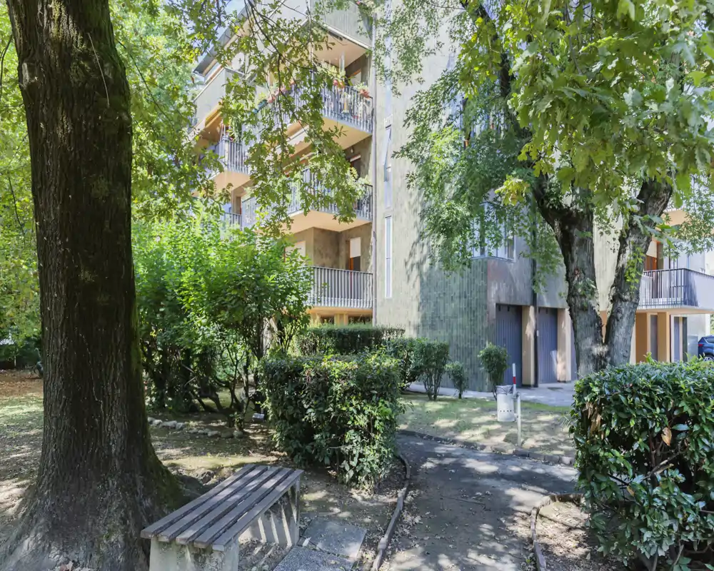 Patio comunitario soleado con árboles maduros, setos recortados y un banco: área exterior tranquila frente a un edificio de apartamentos.