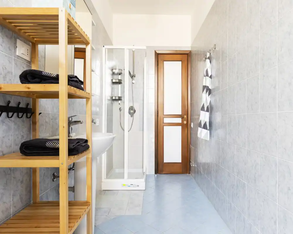 Baño luminoso y moderno con cabina de ducha de cristal, lavabo y estantería de madera para las toallas; azulejos claros y luz natural crean un ambiente fresco y limpio.