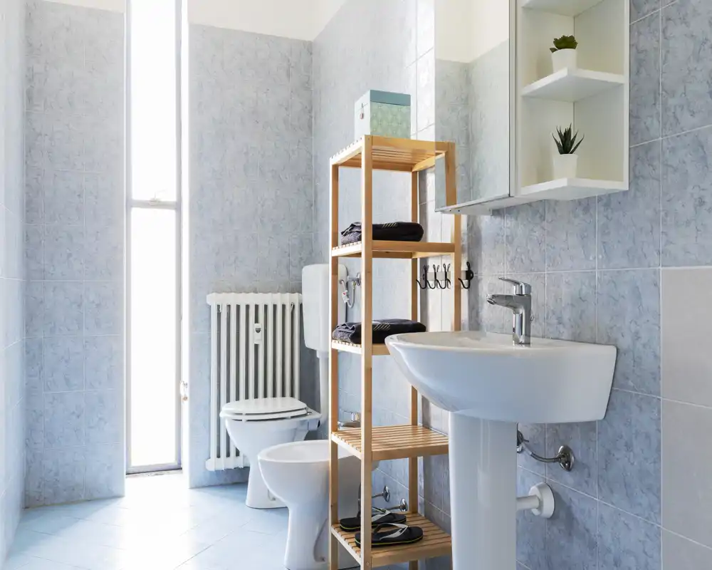 Baño moderno y luminoso con lavabo de pedestal, inodoro, bidé y estantería de madera; azulejos azul claro y una ventana estrecha aportan abundante luz natural.