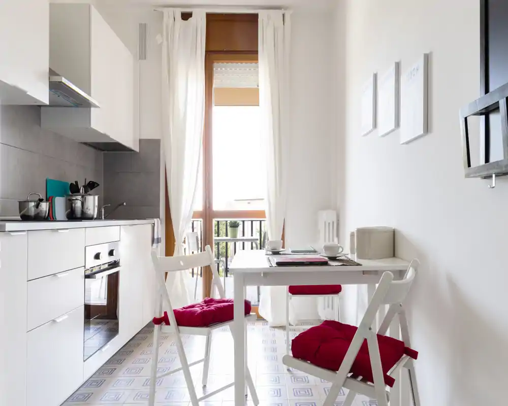 Cocinita luminosa y moderna con una pequeña mesa, sillas con cojines rojos, luz natural desde la puerta al balcón y mobiliario blanco y minimalista.