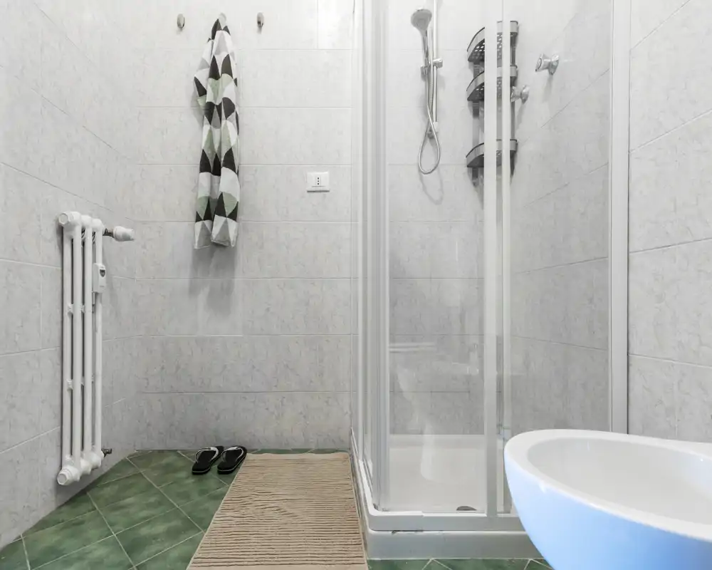 Baño limpio y moderno con mampara de ducha de vidrio, ducha de mano, radiador de pared y lavabo; azulejos grises y suelo verde que crean un espacio funcional.