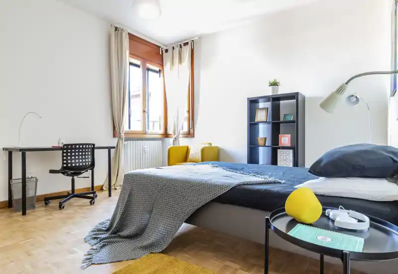 Bright, modern bedroom with a neatly made double bed, bedside table and desk by the window. Soft textiles and a yellow accent armchair give the room a warm, welcoming feel—ideal for rental listings.