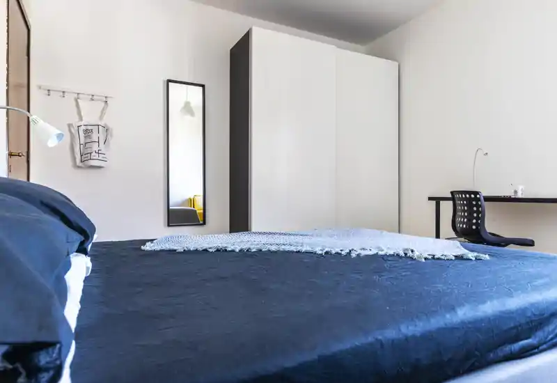 Bright, modern bedroom with a large bed in the foreground, simple wardrobe, full-length mirror and a small desk. Clean linens and a decorative throw give a neat, welcoming look ideal for rental photos.