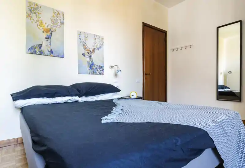 Bright, tidy bedroom with a large double bed dressed in dark bedding and a patterned throw. Two whimsical deer paintings add color above the headboard; a full-length mirror and wooden door complete the simple, modern décor.