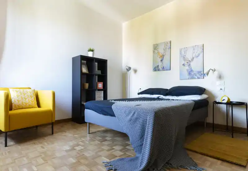 Bright, stylish bedroom with a neatly made double bed, coordinated pillows and a textured throw. The room features a bold yellow armchair, shelving for storage and tasteful wall art for a welcoming atmosphere.