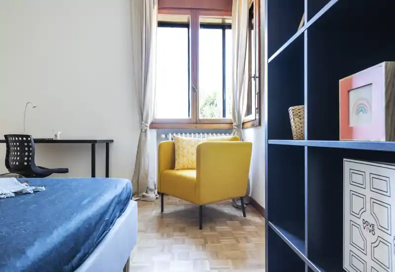 Bright, well-staged bedroom corner with a visible bed in the foreground, a cozy yellow armchair by a large window and shelving to the right — ideal for rental photos showcasing natural light and comfortable seating.