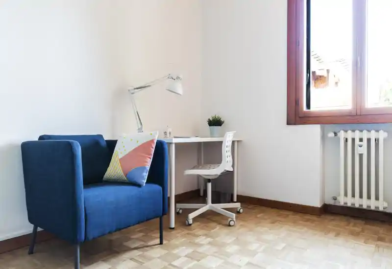 Bright, modern living corner with a blue armchair, small study desk and natural light from a large window — comfortable and stylish for relaxing or working.