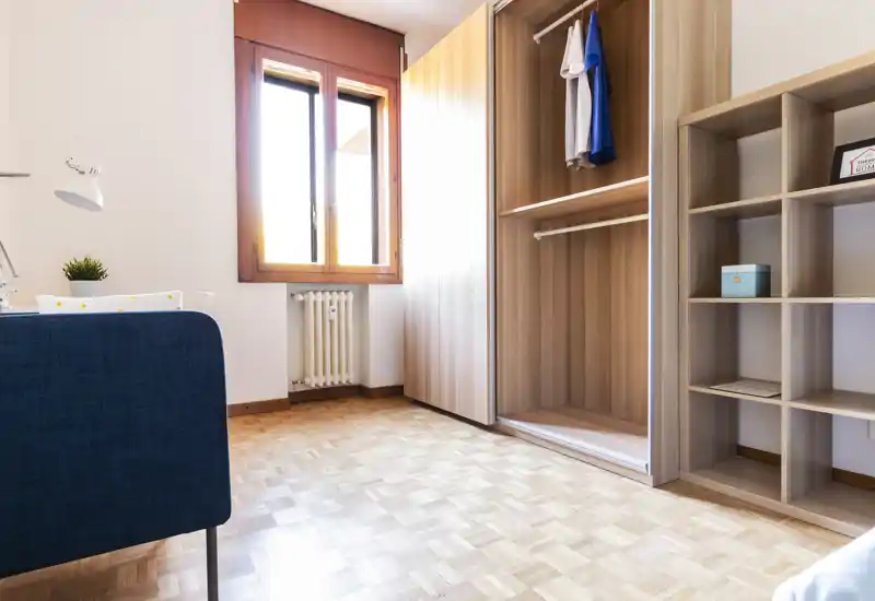 Small, bright bedroom corner with wooden wardrobe and shelving. Natural light from the window highlights the parquet floor and clean, minimal furnishings—ideal for a single occupant.